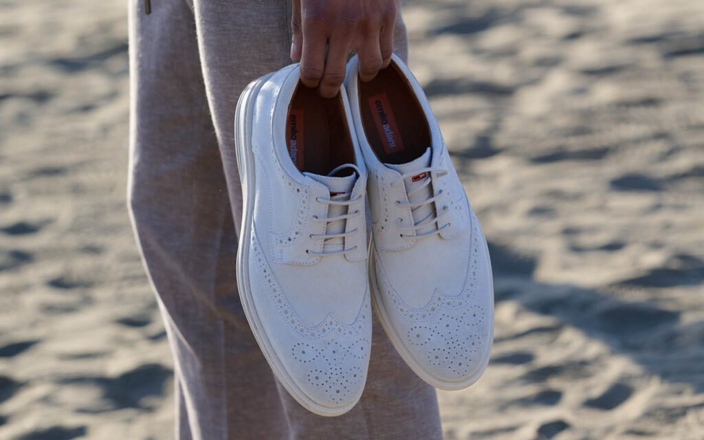 Eine Person hält ein Paar creme Business-Sneaker mit Lochmuster in der Hand, im Hintergrund ist sandiger Untergrund zu sehen.