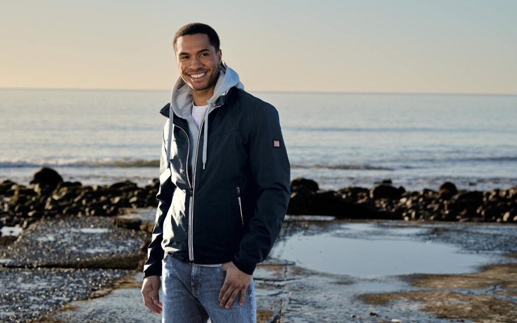 Das Model trägt am Strand eine blaue Blouson Jacke von emilio adani. Sportlich kombiniert mit einem grauen Sweatshirt. 