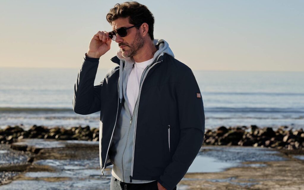 Mann trägt eine dunkle emilio adani Jacke über einem grauen Hoodie am Strand, kombiniert im lässigen Layering-Stil.