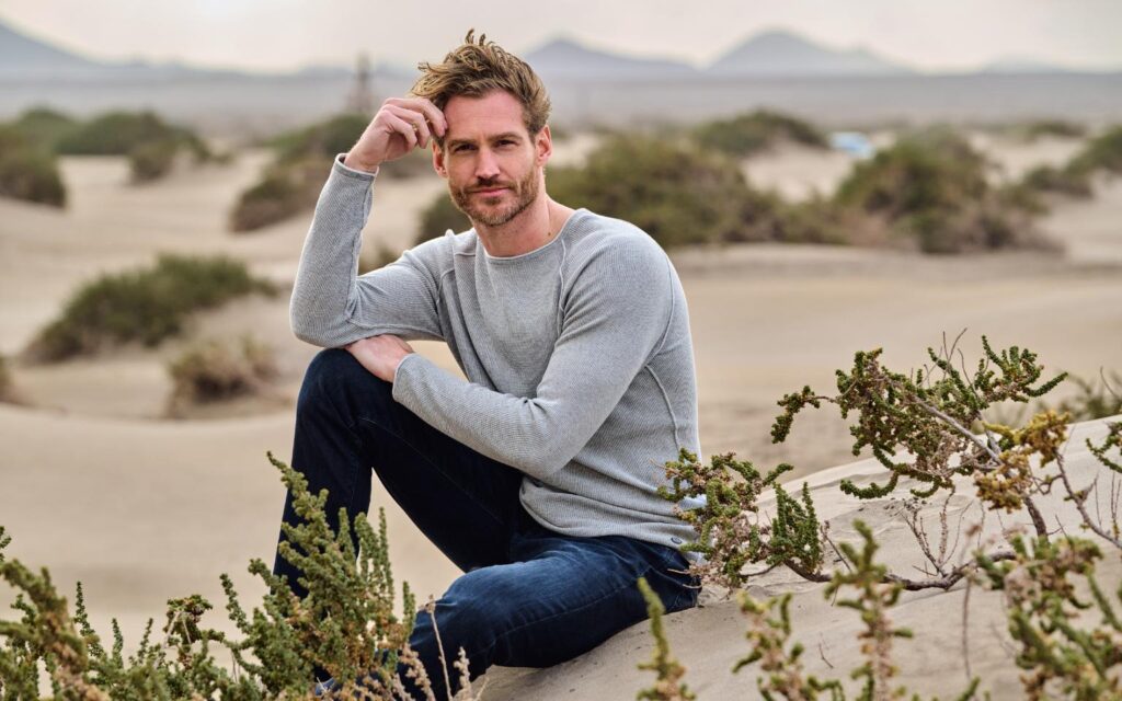 Ein Mann sitzt entspannt in einer sandigen Landschaft und trägt einen grauen engbers Pullover mit dunkler Jeans. 