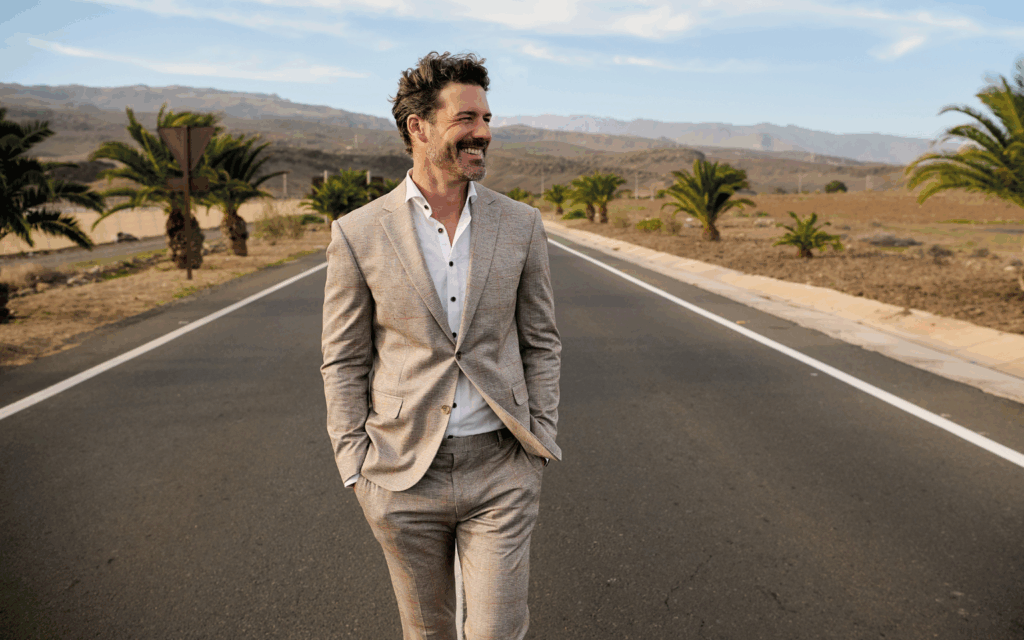 Ein Mann läuft entspannt über eine sonnige Straße in mediterraner Landschaft. Er trägt einen hellen, sommerlichen Anzug mit weißem Hemd und verkörpert einen lässig-eleganten Look.
