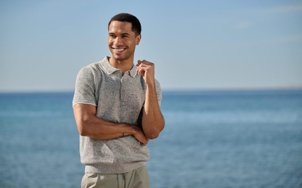 Ein lächelnder Mann trägt ein hellgrünes Poloshirt von emilio adani und steht entspannt vor dem Meer unter blauem Himmel.