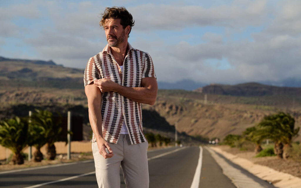 Ein Mann trägt ein gemustertes Kurzarmhemd und eine helle Hose und steht auf einer Straße in einer sonnigen Landschaft mit Bergen und Palmen im Hintergrund.