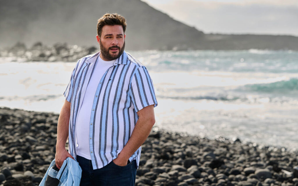 Sommerliches Herren-Outfit mit Hemd Person trägt kurzärmeliges, gestreiftes engbers Hemd über weißem T‑Shirt an einer steinigen Küste.