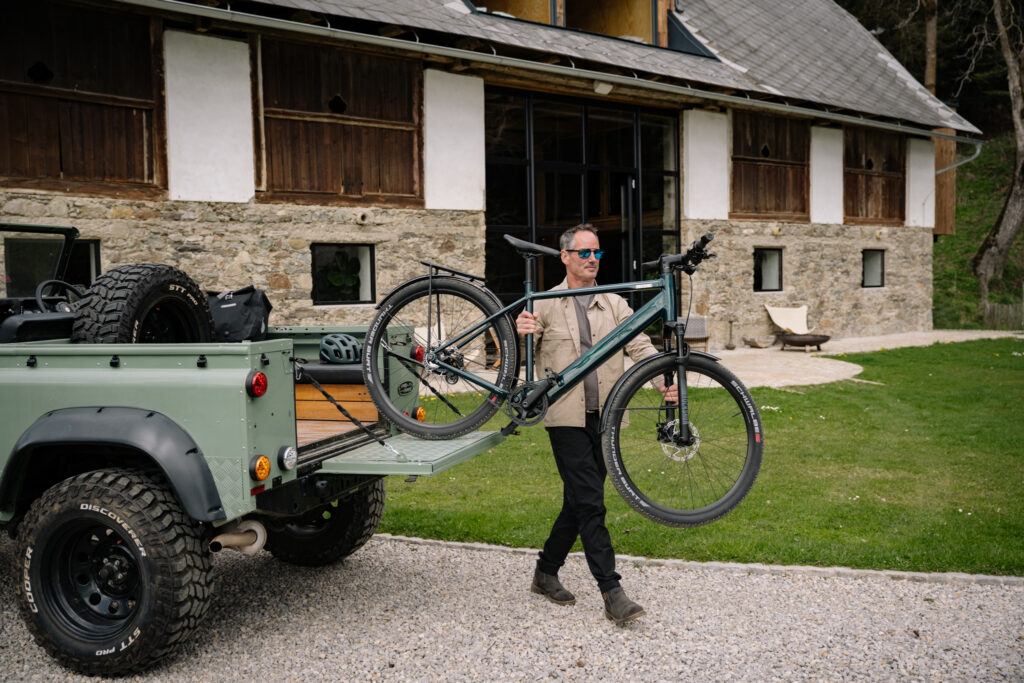 Mit dem coboc E-Bike ins Abenteuer Ein Mann lädt sein coboc E-Bike von einem Geländewagen vor einem rustikalen Landhaus aus – bereit für eine Tour in der Natur.