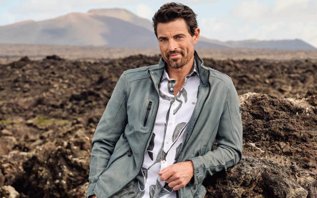 Mann in Lederjacke von engbers lehnt an Felsen in karger Landschaft