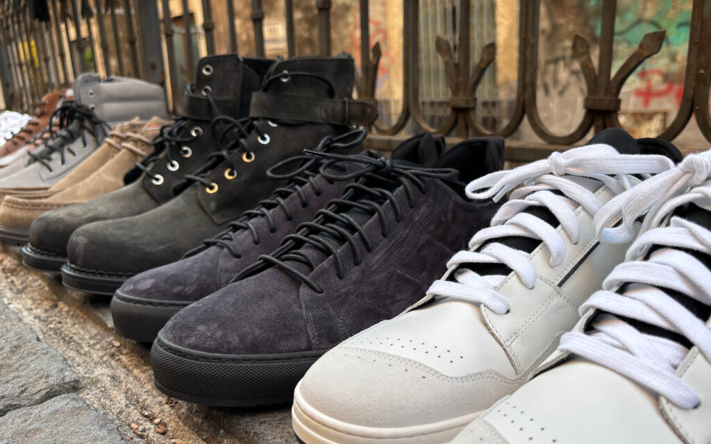 Mehrere Herren-Sneaker und Boots in verschiedenen Farben stehen nebeneinander auf einem Steinboden Weiße Sneaker sowie Boots und Schnürschuhe in Schwarz-, Grau- und Beigetönen sind auf einem gepflasterten Untergrund vor einem Metallgeländer aufgereiht.