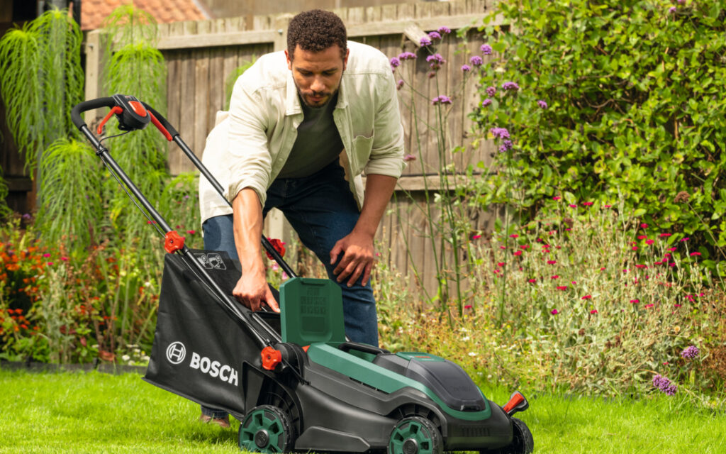 Eine Person setzt den Grasfangkorb in einen elektrischen Rasenmäher der Marke Bosch ein. Der Rasenmäher steht auf einer grünen Rasenfläche in einem Garten mit Blumen, Sträuchern und einem Holzzaun im Hintergrund.