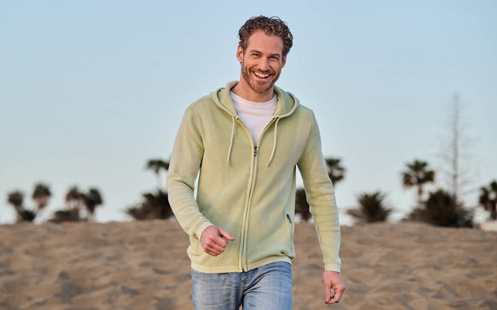 Ein lächelnder Mann geht am Strand entlang. Er trägt einen hellgrünen Strick-Hoodie mit Reißverschluss, ein weißes T-Shirt und eine helle Jeans. Im Hintergrund sind Sand, Palmen und ein heller Himmel zu sehen.