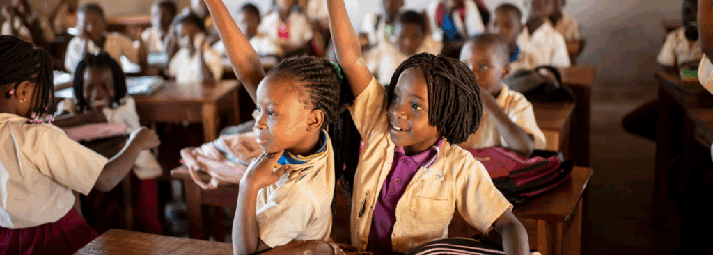 Unterrichtssituation im Klassenzimmer Mehrere Kinder sitzen in einem Klassenraum an Holztischen, einige melden sich, während andere mit Schulmaterialien und Heften arbeiten.