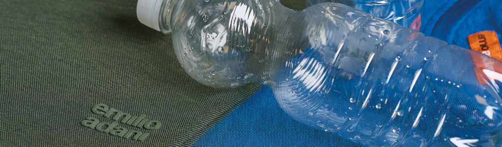 Recycelte Materialien: Detailansicht nachhaltiger emilio adani T-Shirts Nahaufnahme eines grünen und eines blauen T-Shirts aus strukturiertem Stoff. Darauf liegen mehrere leere, transparente Plastikflaschen.