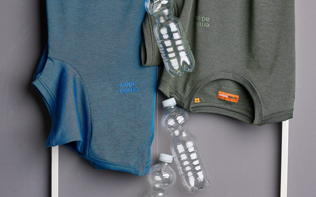 Nachhaltige Mode: T-Shirts aus recyceltem Material bei engbers Zwei gefaltete T-Shirts in Blau und Grün liegen nebeneinander auf einer weißen Fläche. Dazwischen befinden sich mehrere leere Plastikflaschen, die auf das Thema Recycling und nachhaltige Materialverwendung in der Mode hinweisen.