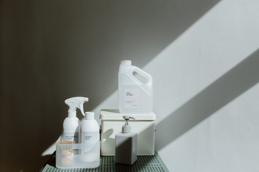 Waschmittel Minimalistische Szene mit weißen Wasch- und Reinigungsprodukten auf einem Tisch. Sichtbar sind ein Flüssigwaschmittel, ein Weichspüler, ein Reiniger und ein Seifenspender. Natürliches Licht und starke Schatten sorgen für eine moderne, ruhige Atmosphäre.