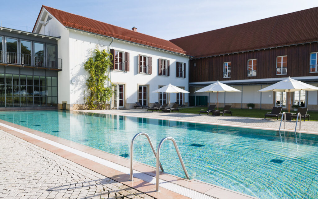 Außenpool und Hotelanlage im Gräflichen Park Großer Außenpool vor einer Hotelanlage. Am Beckenrand stehen Liegen und Sonnenschirme auf einer sonnigen Terrasse.