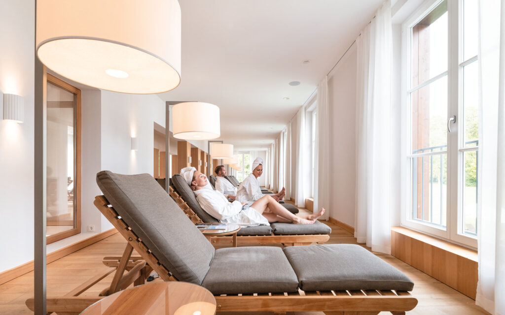Ruhebereich im Spa- und Wellnessbereich Mehrere Personen entspannen auf Liegen in einem hellen Wellnessbereich mit großen Fenstern und Holzfußboden.