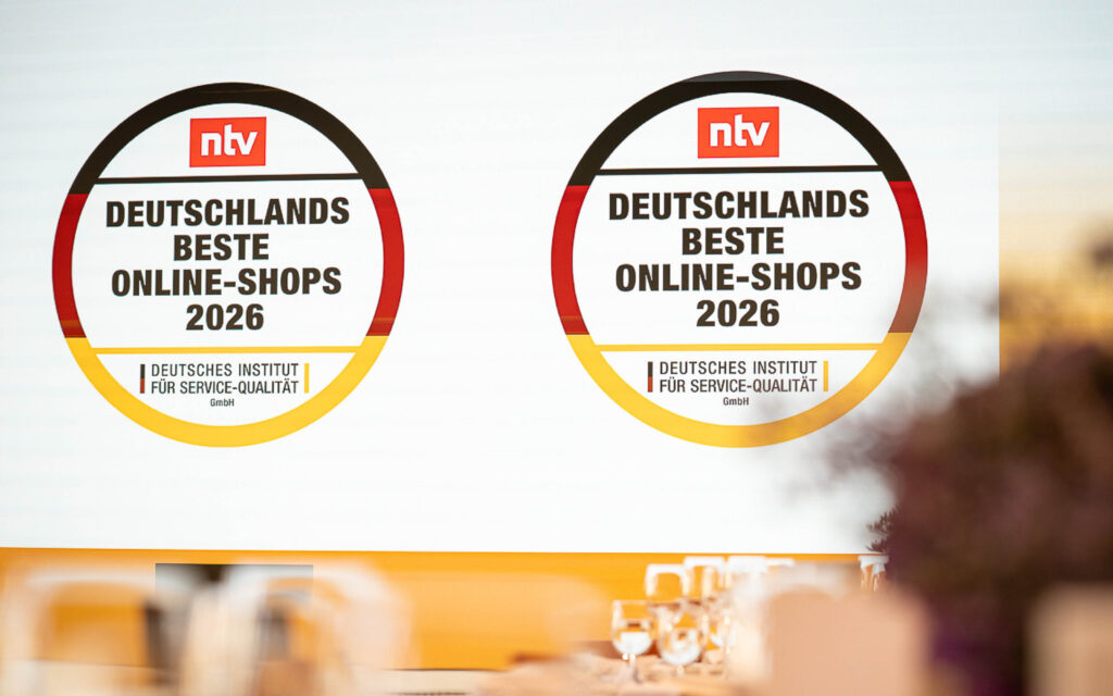 Auszeichnung „Deutschlands Beste Online-Shops 2026“ Zwei Auszeichnungen „Deutschlands Beste Online-Shops 2026“ auf einer Präsentationsfläche. Im Vordergrund sind unscharfe Gläser zu sehen.